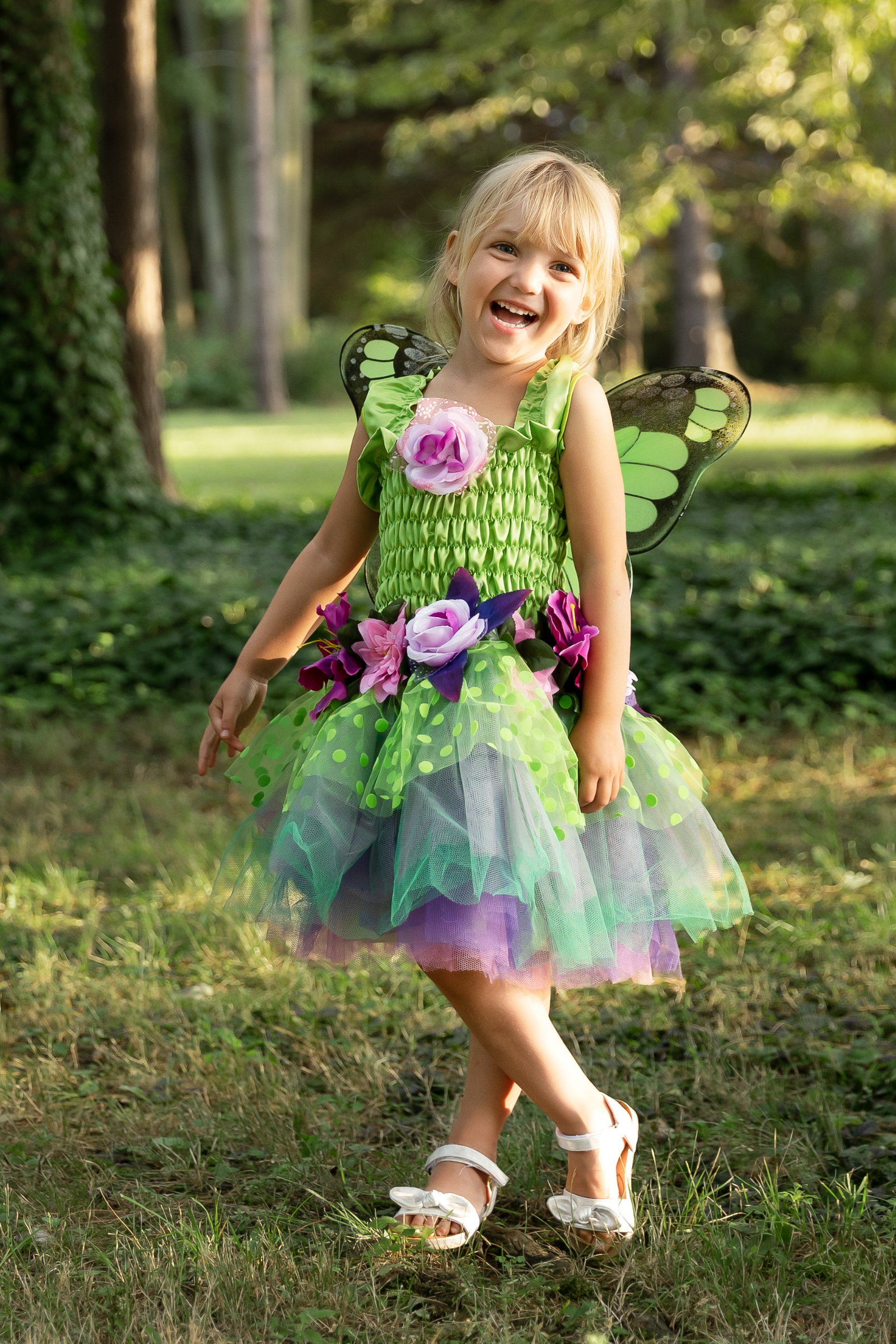 A young child dressed in a green and purple fairy costume with a tulle skirt and detachable wings, standing in a garden setting.